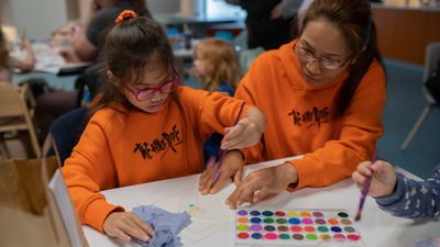 A mother and child, both wearing bright orange sweatshirts, are sitting at a table painting a picture together. In front of them is a box of paints.