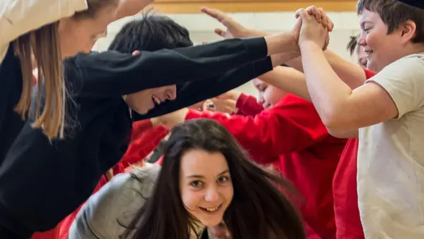 A photograph of a group of children ceilidh dancing. Two rows of children are standing opposite each other, arms raised and hands touching to create an arch. A pair of children holding hands is dancing through the created arch, looking forwards at the camera.