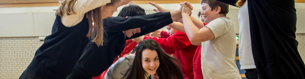 A photograph of a group of children ceilidh dancing. Two rows of children are standing opposite each other, arms raised and hands touching to create an arch. A pair of children holding hands is dancing through the created arch, looking forwards at the camera.