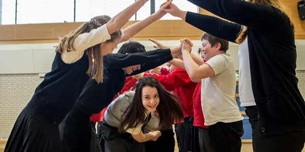 A photograph of a group of children ceilidh dancing. Two rows of children are standing opposite each other, arms raised and hands touching to create an arch. A pair of children holding hands is dancing through the created arch, looking forwards at the camera.