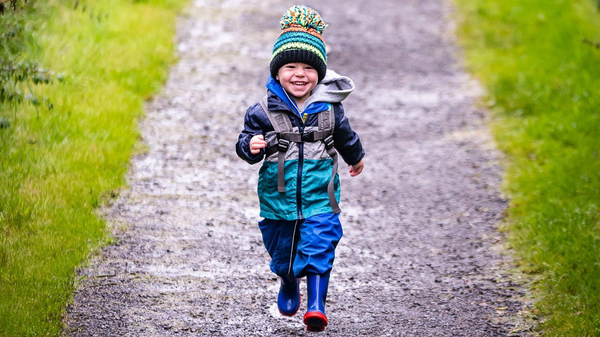 A small child dressed in a waterproof suit, wellington boots and a bobble hat is running along a path with grass on either side. The child is smiling.