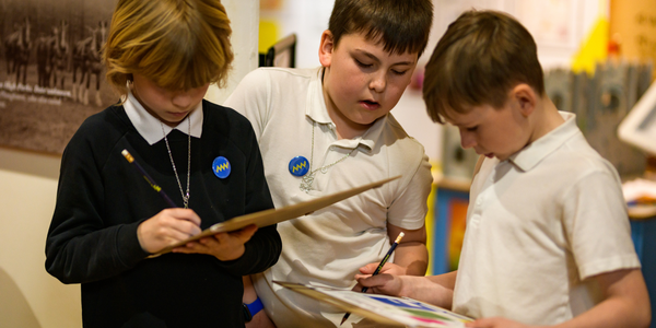 Three school children. The one on the left wearing a navy jumper and writing on a clipboard. Next to him on the right are two children in white polo shirts, one of which is also holding a clipboard. The third child is watching them write.
