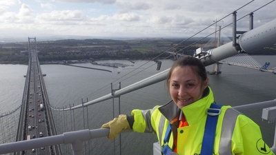 A woman in a yellow high vis vest above the Forth Road Bridge. She is smiling at the camera.