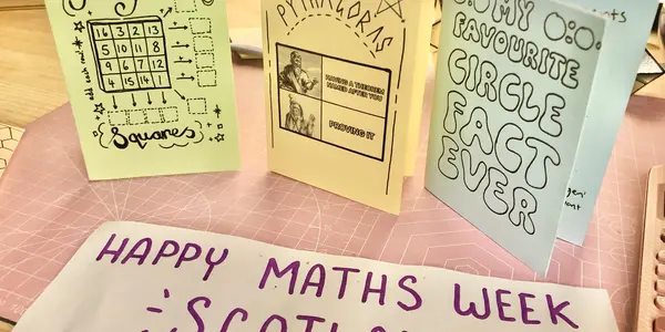Three folded paper maths zines standing upright on a desk. On the desk is a piece of paper that says Happy Maths Week Scotland.