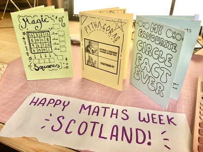 Three folded paper maths zines standing upright on a desk. On the desk is a piece of paper that says Happy Maths Week Scotland.