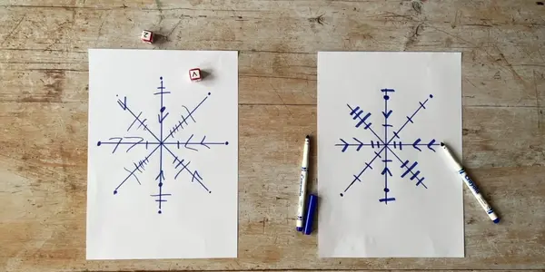 Two sheets of white paper with blue markers and dice. Each sheet has a drawing of a snowflake on it.