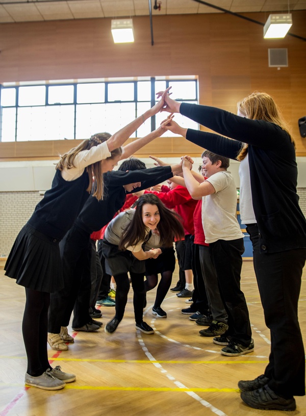 A photograph of a group of children ceilidh dancing. Two rows of children are standing opposite each other, arms raised and hands touching to create an arch. A pair of children holding hands is dancing through the created arch, looking forwards at the camera.