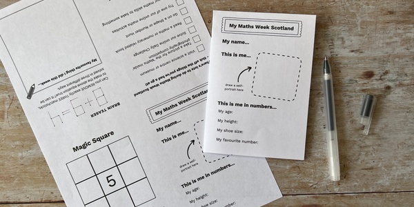 Two sheets of paper on a wooden desk top. One sheet is flat, the other is folded up. The sheets show the content of the My Maths Week Scotland activity booklets. Next to the sheets is an open pen.